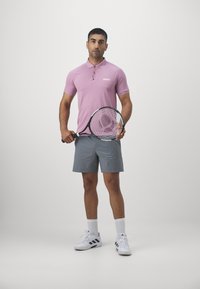 Camisa de polo rosa con cuello y logo, pantalones cortos grises, sosteniendo una raqueta de tenis, vistiendo zapatillas deportivas y calcetines blancos, de pie sobre un fondo liso.