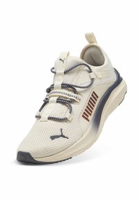 Zapatilla de malla beige con acentos en azul oscuro, superficie texturizada y cordones elásticos. Presenta un destacado logotipo de "PUMA" en naranja.