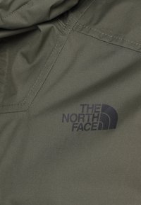 Olivengrønt stof med en glat tekstur, der har et sort logo af "THE NORTH FACE" på nederste højre side. Sømme detaljer er synlige.