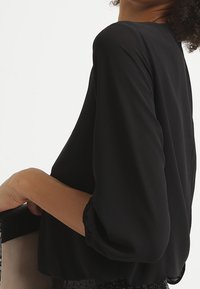 Nainen, jolla on päällään musta pitkahihaista puseroa ja puffihihnoja, pitää beigen ja mustan käsilaukun valkoista taustaa vasten.