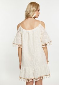 Bijela kratka haljina s čipkastim dizajnom, off-shoulder rukavima i resicama na rubu. Rašireni rukavi dodaju teksturu i vizualni interes.