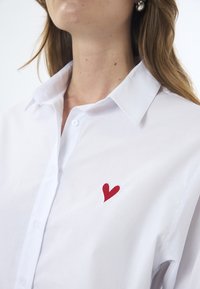 Weißes Baumwollhemd mit Knöpfen und einem spitzen Kragen. Verfügt über ein rotes gesticktes Herz im linken Brustbereich. Glatte Textur.