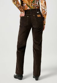 Bruna kordflortbyxor med vertikala ribbor, utrustade med två bakfickor och en läderlogg, tillsammans med svarta skor.