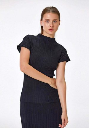 Femme à la peau claire et aux yeux bleus portant un top noir côtelé à manches courtes et une jupe assortie, posant devant un fond blanc uni.