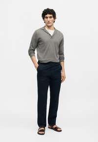 Homme debout portant un polo gris à manches longues, pantalon bleu marine et sandales marron, une main dans la poche, fond neutre.