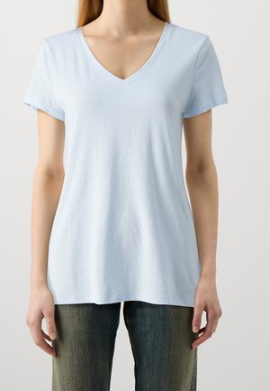 Ung person iført lyseblå V-hals T-shirt og slidte blå jeans, stående foran en ensfarvet hvid baggrund.