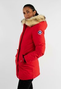 Parka d'hiver rouge avec capuche en fausse fourrure. Comprend des fermetures à bouton, des poches latérales et un patch sur la manche gauche. Texture lisse et coupe au niveau des hanches.