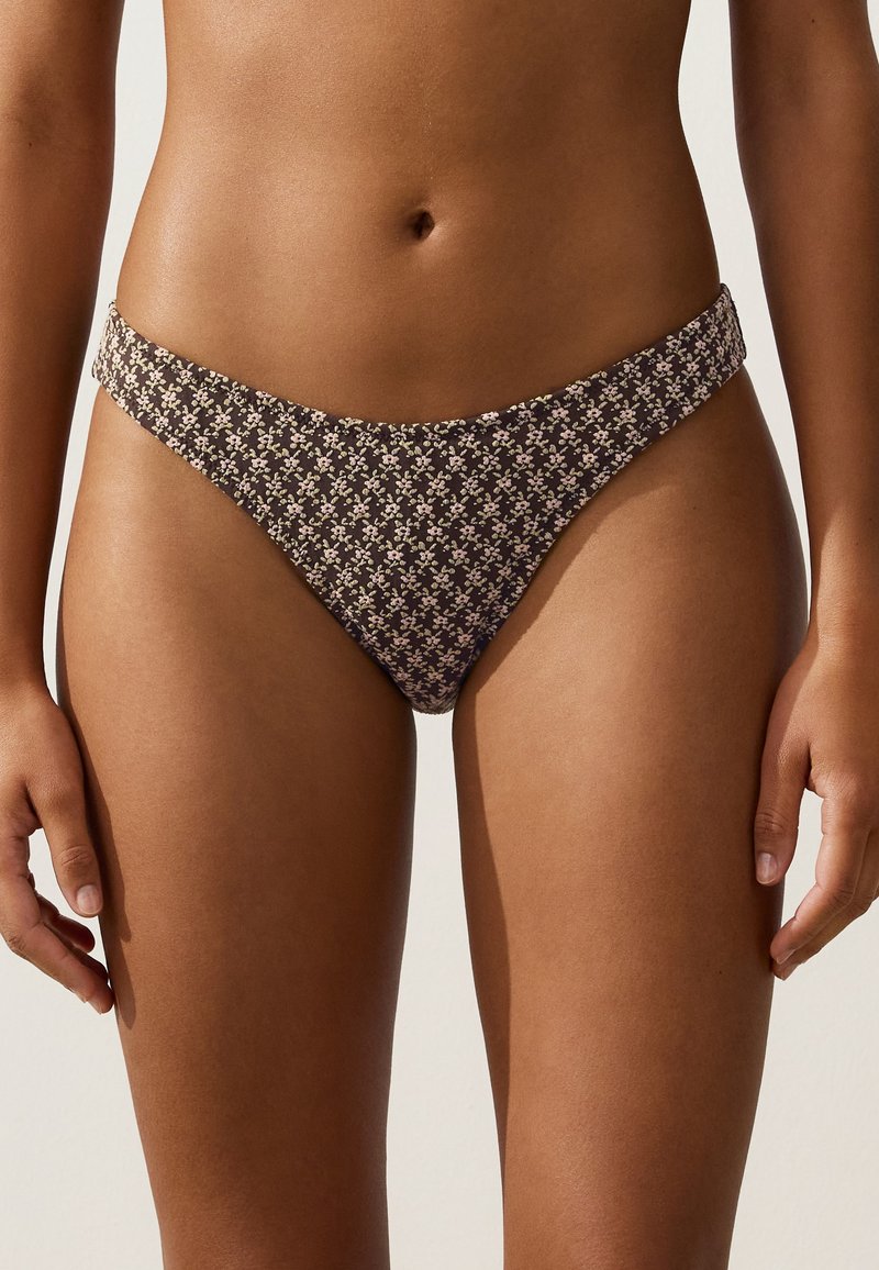 Buste féminin portant un bas de bikini échancré à petit motif fleuri sur fond sombre, devant un fond uni clair.