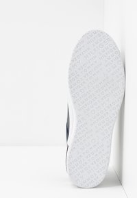 Semelle de basket blanche avec texte de logo en relief, tige foncée contrastante, forme épurée et design minimaliste sur fond clair.