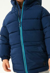 Veste matelassée bleu marine avec une fermeture éclair turquoise, des poignets élastiques et un logo souriant. Équipée d'une capuche et de deux poches latérales.