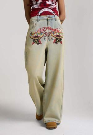 Weit geschnittene hellblaue Jeans mit bunten Ed Hardy Stier- und Sternmotiven auf den Gesäßtaschen, kombiniert mit braunen Schuhen und einem roten Grafiktop.