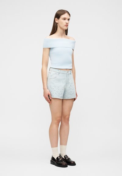 Γυναίκα με ανοιχτό γαλάζιο τοπ με ακάλυπτους ώμους, ανοιχτόχρωμα τζιν σορτς με πιτσιλιές από μπογιά, κρέμ κάλτσες και μαύρες μοκασίνες, στέκεται μπροστά σε απλό φόντο.