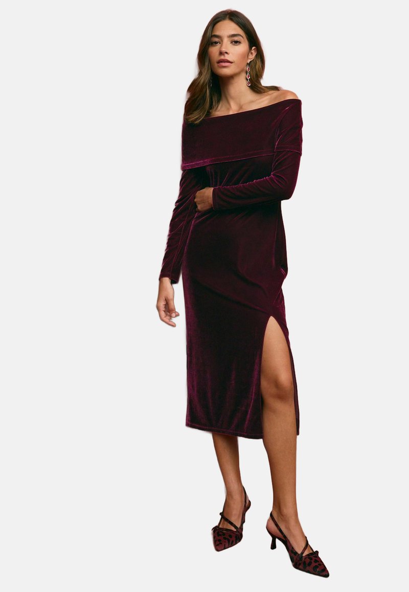Burgundyröd sammetsklänning med off-shoulder-axlar och långa ärmar, med en sidoslits och en figurnära design, i kombination med spetsiga klackar.
