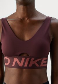 Közelkép egy személyről, aki bordó sportmelltartót visel, kulcslyuk kivágással és egy nagy betűkkel „NIKE” feliratozott elasztikus pánttal.