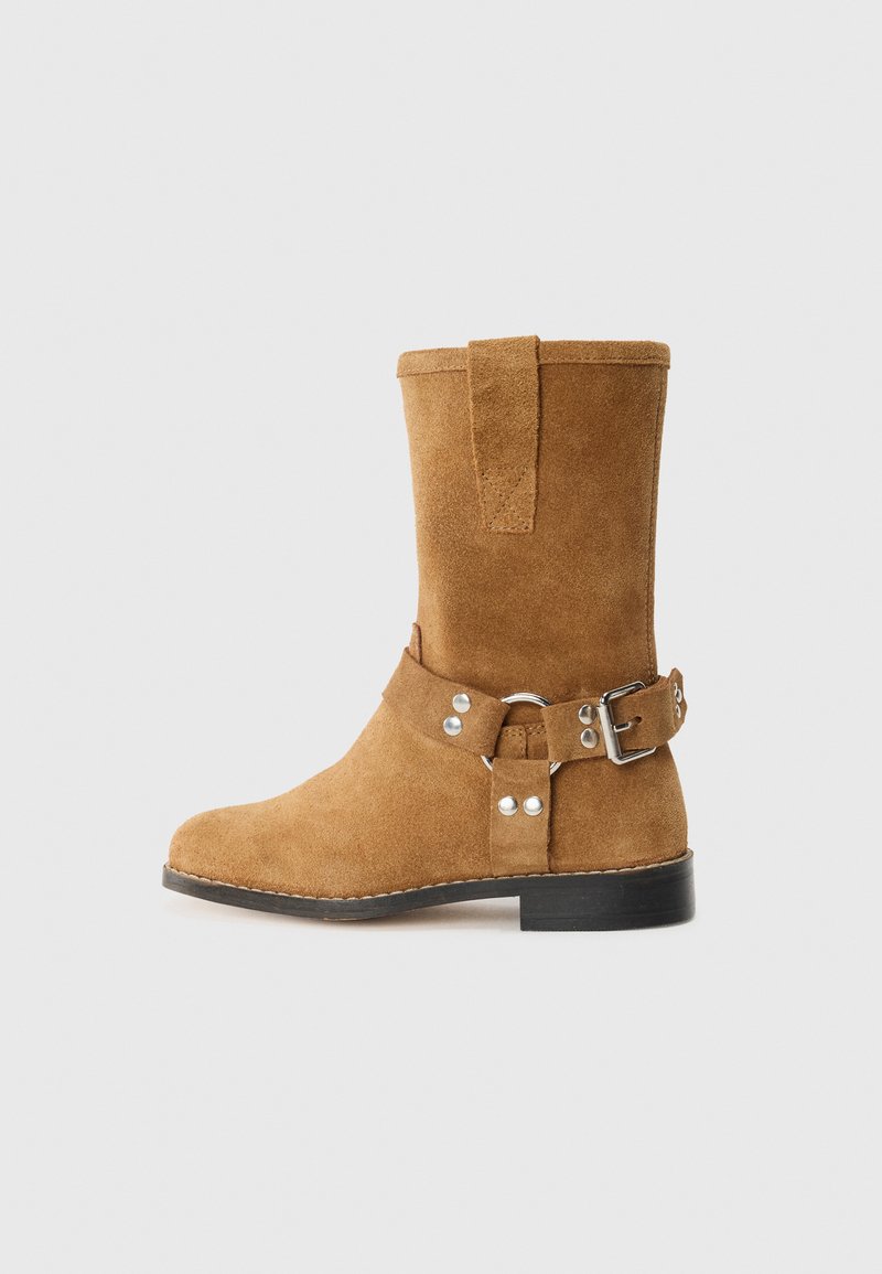 Bota de tornozelo em camurça bege com ponta arredondada, uma tira com fivela ao redor do tornozelo e uma sola preta plana. Inclui uma alça na parte de trás para facilitar o calçar.