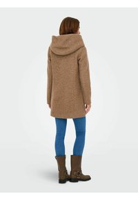 Bruine textuurjasje met een grote capuchon, lange mouwen en een rechte snit, gecombineerd met blauwe jeans en bruine laarzen.