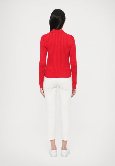 Rdeč pulover z vzorcem pletenja, ovratnik, oprijet kroj, rebrasti robovi pri zapestjih in spodnjem robu, kombiniran z belimi ohlapnimi hlačami in belimi supergami.