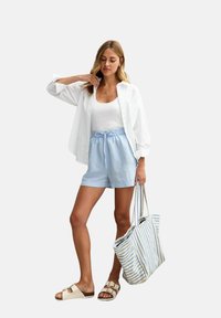 Camicia bianca a bottone sopra una canotta bianca, pantaloni corti azzurri con cintura elastica, abbinati a una borsa a righe e sandali.