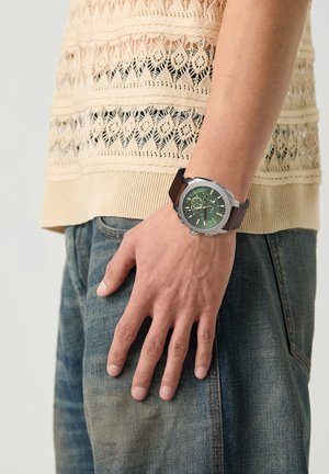 Persona que lleva un reloj de pulsera con esfera verde y correa de cuero marrón, top beige de encaje tejido y jeans vaqueros azules, brazo relajado a su lado.