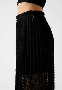 Gonna plissettata nera in pizzo, con un design strutturato e una vita aderente con un dettaglio accentato in argento. Pannello inferiore semitrasparente.