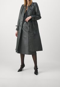 Ciemnoszara skórzana trench coat z podwójnym zapięciem, shoulder epauletami i paskiem w talii, zestawiona z dopasowaną skórzaną spódnicą i pończoszkami w kropki.