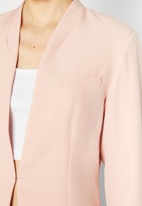 Žena v světle růžovém saku přes bílý žebrovaný crop top, který odhaluje horní část hrudníku a oblast u krku.