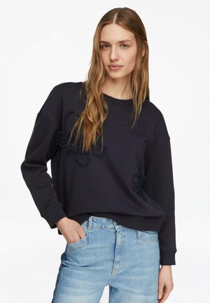 Kobieta o długich, jasnobrązowych włosach, ubrana w luźną czarną bluzę i jasnoniebieskie dżinsy z wysokim stanem, stojąca z jedną ręką w kieszeni.