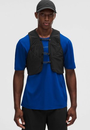 Junger Mann mit schwarzer Kappe, blauem kurzärmeligen Hemd und schwarzer Utility-Weste mit Reißverschlusstaschen und verstellbaren Verschlüssen.