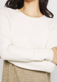 Pull en maille couleur crème avec un col rond et des manches longues, présentant une texture douce et un design de manches raglan.