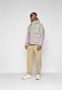 Hellgraue wasserdichte Jacke mit einer lavendelfarbenen Fronttasche, mehreren Taschen und Kapuze, kombiniert mit beigen Cargo-Hosen und weißen Sneakers.
