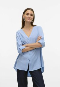 Lichtblauwe, knoopsluiting blouse met een V-hals en omgeslagen mouwen. Heeft een losse pasvorm en een gestructureerde stof. Gedirigeerd met donkere denim.