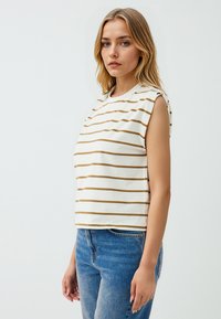 Top senza maniche a strisce bianche con linee orizzontali dorate, scollo rotondo e vestibilità rilassata, abbinato a jeans in denim blu.