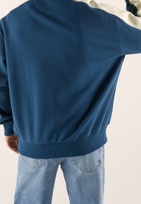 Felpa blu con pannello bianco sulle spalle, polsini e orlo a costine, realizzata in tessuto morbido. Indossata con jeans azzurri chiari, con una vestibilità comoda.