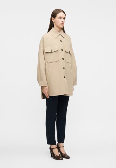 Γυναίκα με μπεζ φαρδιά κουμπωτή παλτό με μεγάλες τσέπες στο στήθος, μαύρο στενό παντελόνι και καφέ ψηλοτάκουνα παπούτσια Mary Jane, που στέκεται μπροστά σε λευκό φόντο.