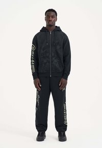 Svart zippered hoodie med grafiska mönster och dragsko, matchad med svarta sweatpants som har drake och texttryck.