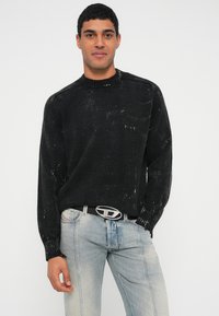 Fekete kötött pulóver texturált felülettel, bordázott kerek nyakkal és hosszú ujjakkal. Világoskék farmernadrággal és ezüst övvel párosítva.