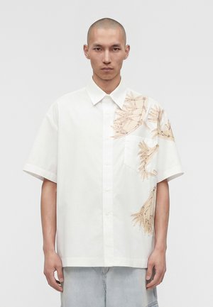 Homme portant une chemise blanche à manches courtes avec des motifs d'ailes d'oiseau beige sur l'épaule droite et la poitrine, associée à un jean bleu clair.