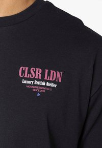 Černá látka zobrazuje růžový a bílý text s nápisem "CLSR LDN Luxury British Atelier Modern Essentials Since 2015" a pod ním malá modrá hvězda.