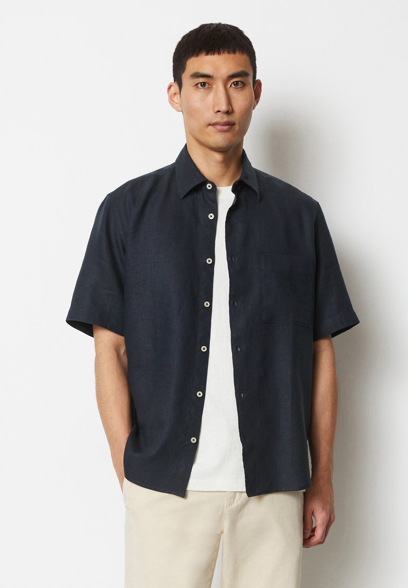 Korte mouwen shirt in donkerblauw, gemaakt van gestructureerde stof met een knoopsluiting aan de voorkant, een enkele borstzak en witte knopen. Draagt over een licht t-shirt.