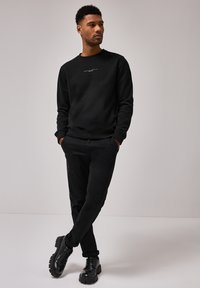 Sort sweatshirt med rund hals og logo, parret med sorte bukser og kraftigt sålede sorte sko. Glat tekstur, tætsiddende design.