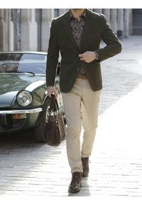 Dark green blazer, patterned shirt, light beige trousers, brown boots, and a brown leather bag. Vintage green car in the background.