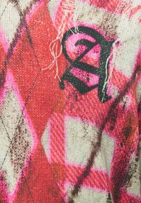 Κοντινή λήψη πλεκτού υφάσματος με κόκκινο, άσπρο και ροζ σχεδιασμό αργκάιλ, με μαύρο κεντημένο στυλιζαρισμένο γράμμα "A" σε υφή μάλλινης επιφάνειας.