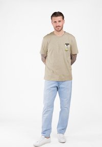 Hombre con brazos tatuados vestido con una camiseta beige que dice "HOPE" y presenta una paloma blanca, jeans de mezclilla claros y zapatillas blancas, de pie frente a un fondo blanco.