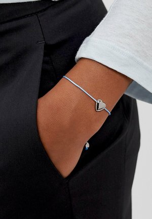 Blaues Schnurarmband mit einem metallischen Herz-Anhänger, getragen an einer Hand, mit sichtbarer Hauttextur und Stoff eines hellen Ärmels.
