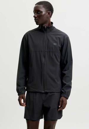 Homme portant une veste noire zippée et un short noir assorti, regardant vers sa gauche sur un fond clair uni.