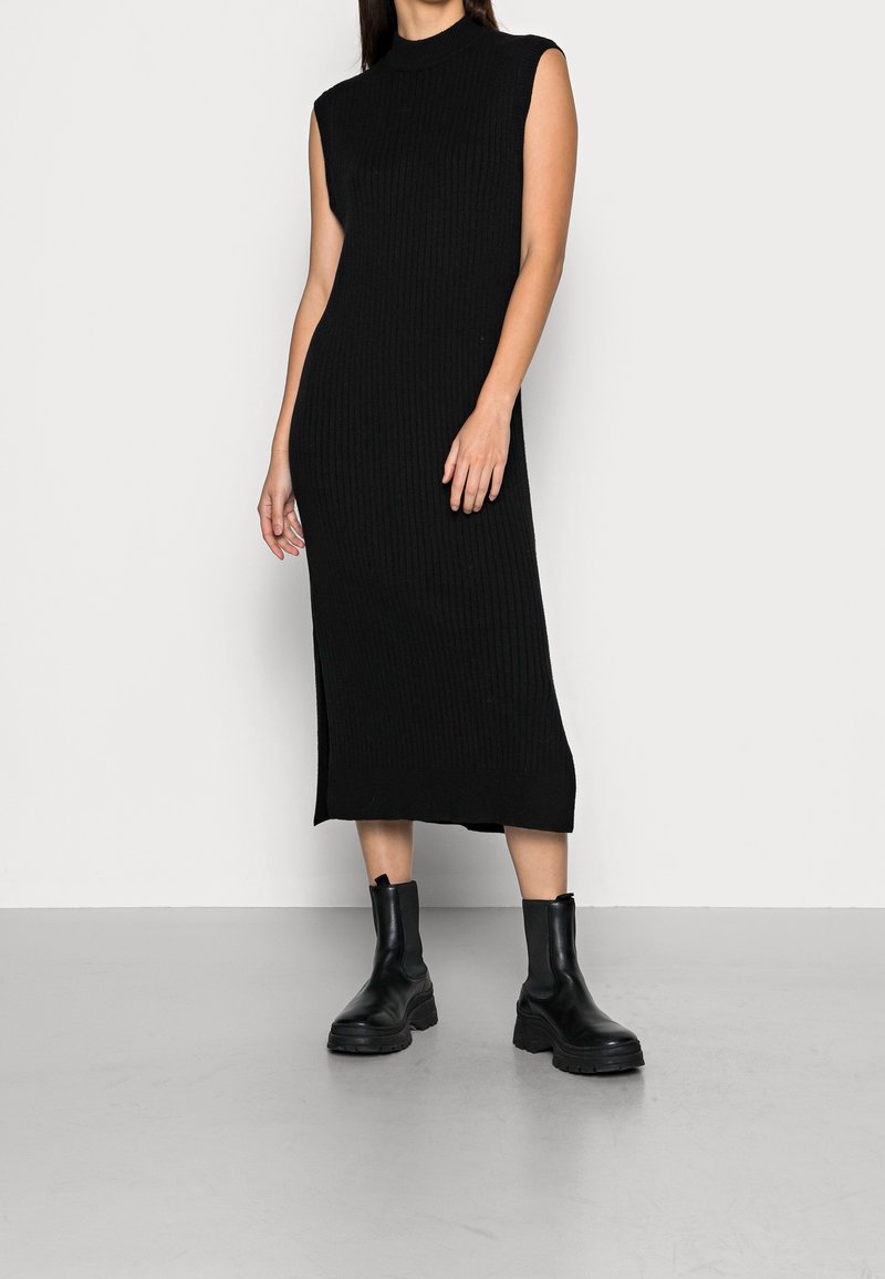 Femme portant une robe midi noire côtelée sans manches associée à des bottines noires épaisses, debout devant un fond clair uni.