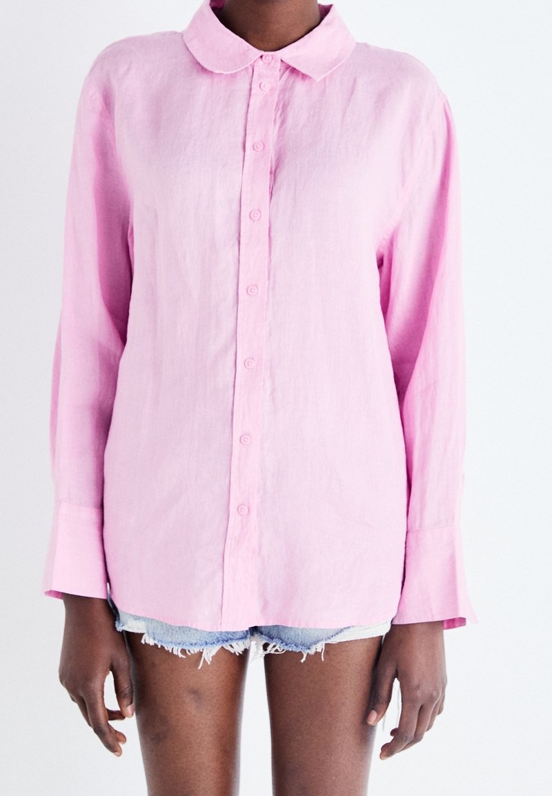 Persona che indossa una camicia rosa a maniche lunghe con colletto arrotondato e pantaloni shorts in denim chiaro sfilacciati contro uno sfondo bianco.
