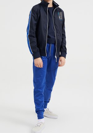 Bambino con giacca blu navy con cerniera e dettagli a strisce blu, pantaloni della tuta blu coordinati, in piedi con scarpe da ginnastica bianche su uno sfondo neutro.