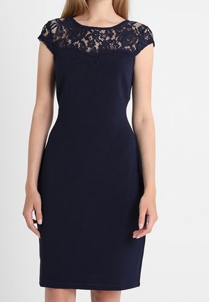 Getailleerde donkerblauwe jurk tot op de knie met kantdetail op de bovenborst en kapmouwen, gedragen door een staande vrouw.