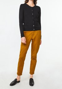 Kvinna i senapsgula byxor, svart knappad cardigan och svarta slip-on-skor, stående mot en enkel vit bakgrund.